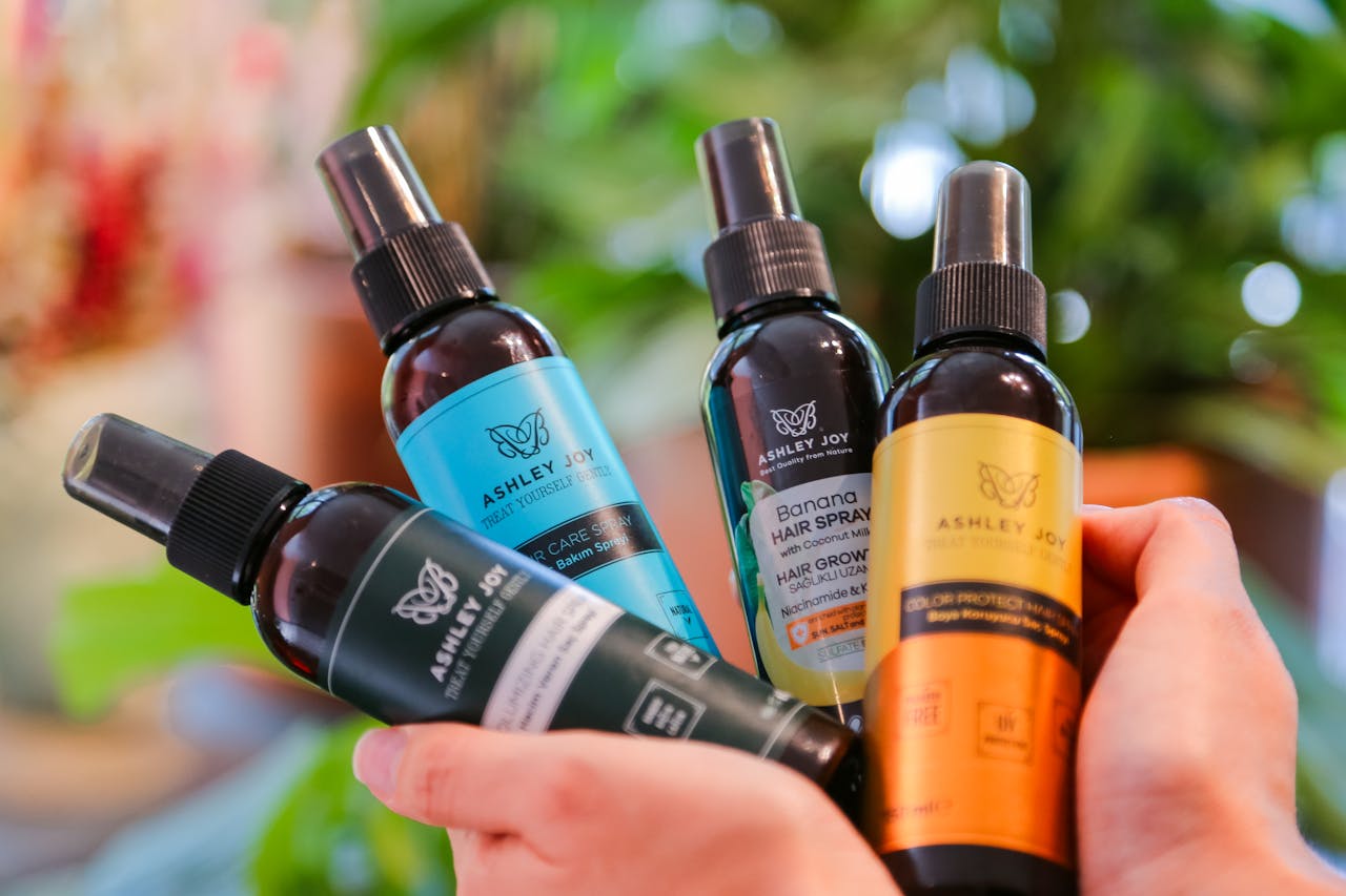 Close-up of hands holding various Ashley Joy hair care bottles with a blurred natural background.